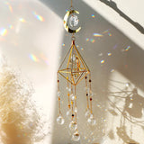 Gold geometric crystal suncatcher casting rainbow light on a pale wall beside dried grasses