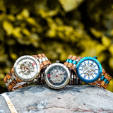 Pair of eco-friendly wooden watches with blue dials on a rock with greenery behind