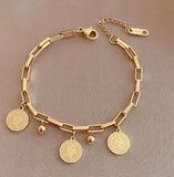 Gold paperclip chain bracelet with three coin charms and bead detail on pink background