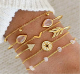 Gold bangle stack with arrow, triangle and bar geometric charms on a pink background