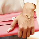 Gold knot bangle with initial charm worn on a wrist beside a pink clutch