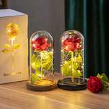 Gold and red foil roses in glass domes with LED lights on wooden bases beside gift box