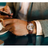 Gold skeleton watch with brown leather strap worn on wrist with a suit sleeve