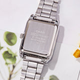 Silver stainless steel tonneau watch with white dial resting beside green leaves