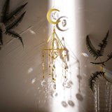 Gold crescent moon suncatcher hanging against a wall with dried leaves and soft shadows