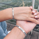 Two hands held together wearing matching silver ID bar bracelets outdoors